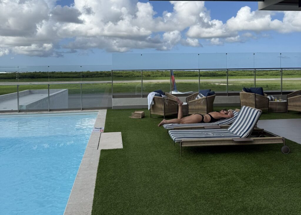 Rooftop pool at the Punta Cana Airport VIP Lounge with lounge chairs, green turf, and a view of the runway under bright blue skies and clouds.