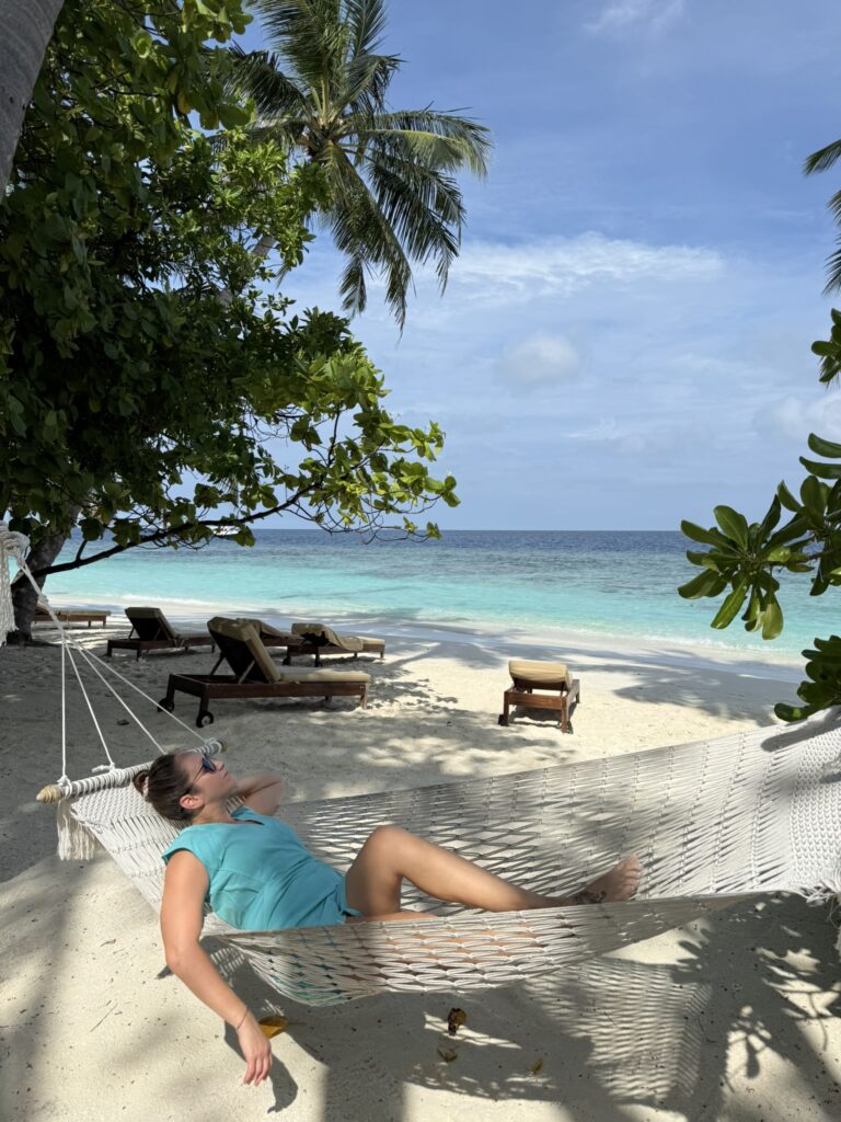Woman relaxing in a white hammock on a sandy beach under palm trees, with lounge chairs and turquoise ocean water in the background.