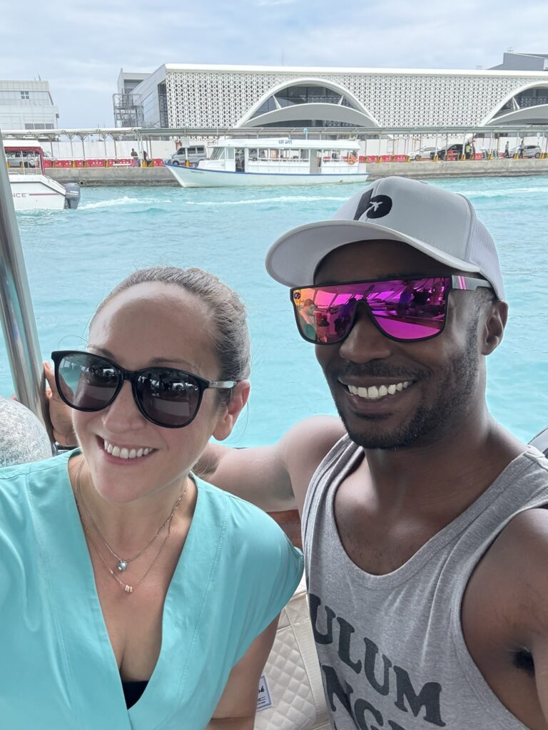 Couple smiling for a selfie on a boat in the Maldives, with bright blue water and the Malé airport terminal visible behind them