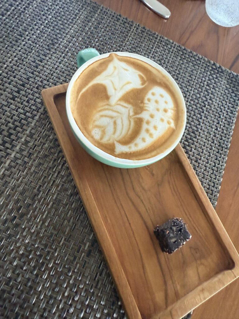Cup of latte with intricate animal-inspired latte art served on a wooden tray, accompanied by a small chocolate brownie on a woven placemat.