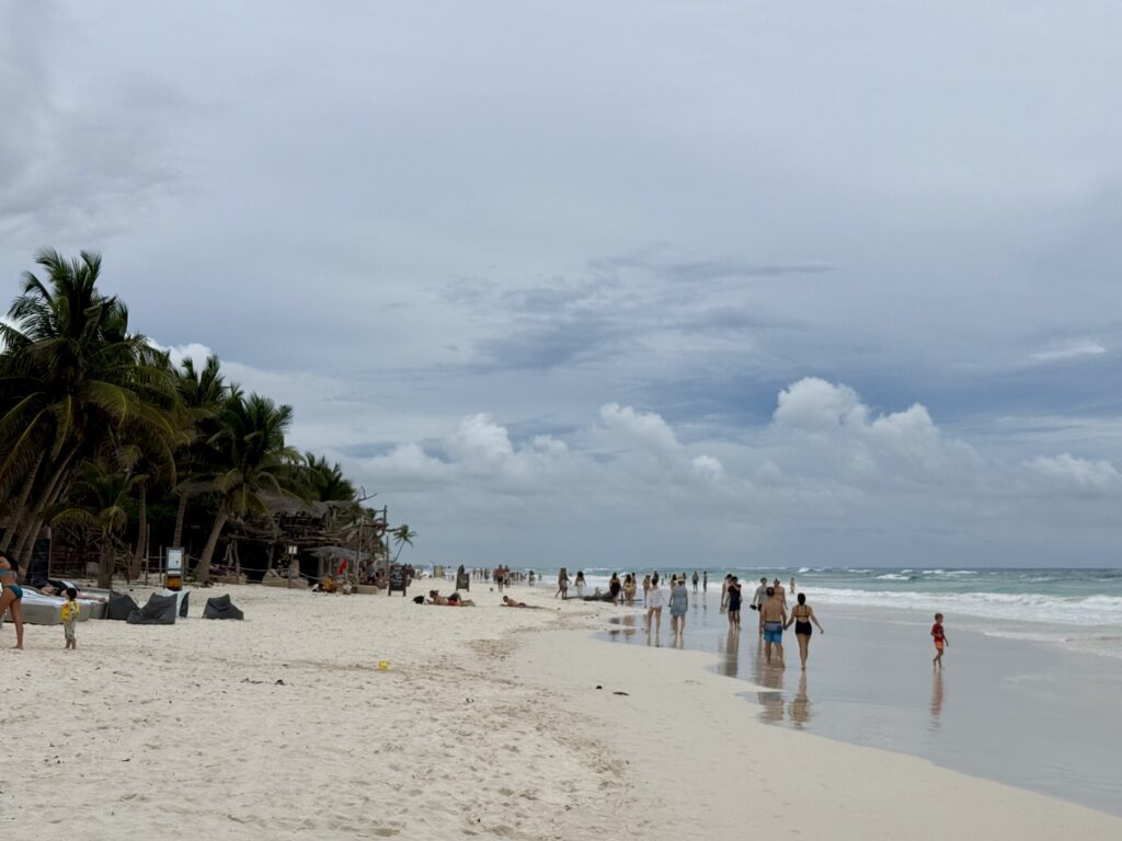 People walking and relaxing along a wide sandy beach in Tulum, Mexico, with gentle waves, turquoise water, and leaning palm trees under an overcast sky.