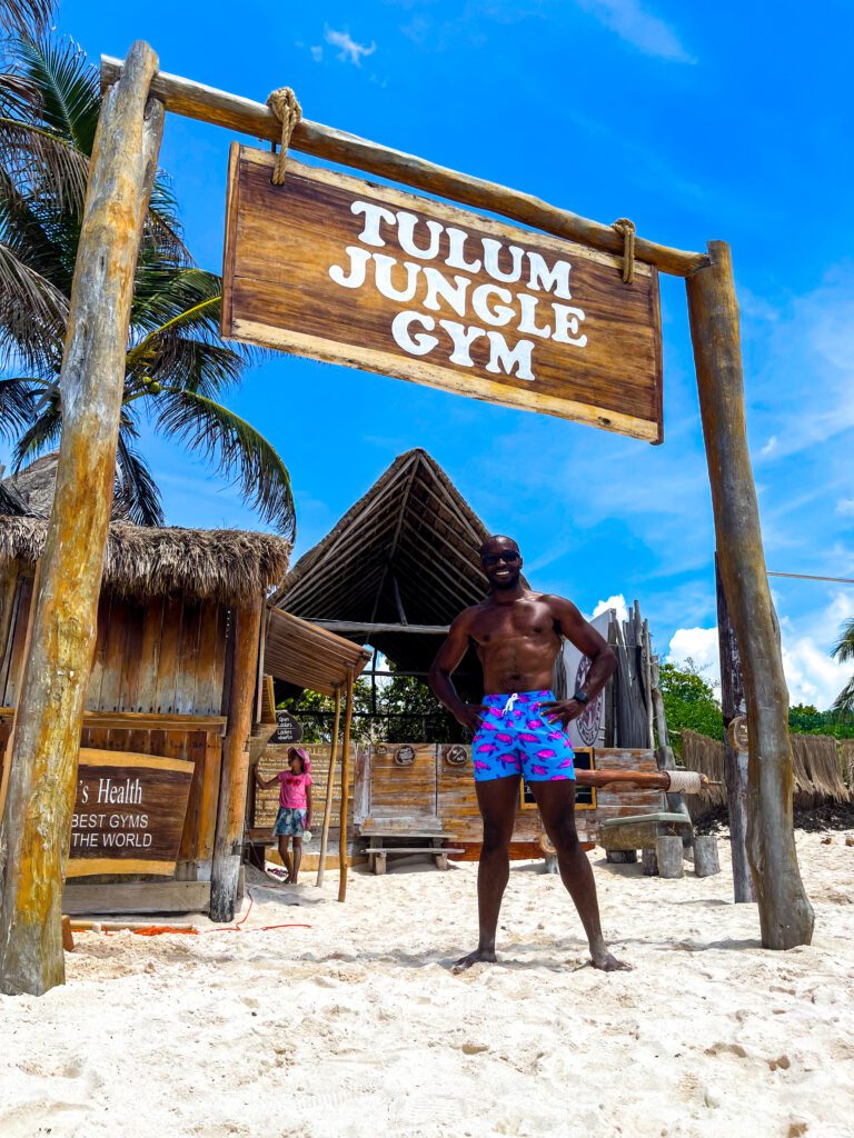 Man standing on a sandy beach beneath the wooden ‘Tulum Jungle Gym’ sign, with rustic beachfront gym structures and palm trees in the background under a bright blue sky