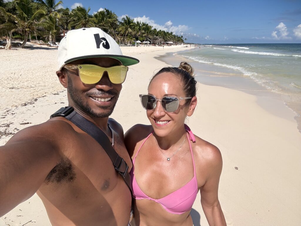 Couple taking a selfie on a sunny beach in Tulum, standing on soft white sand with palm trees and gentle waves in the background