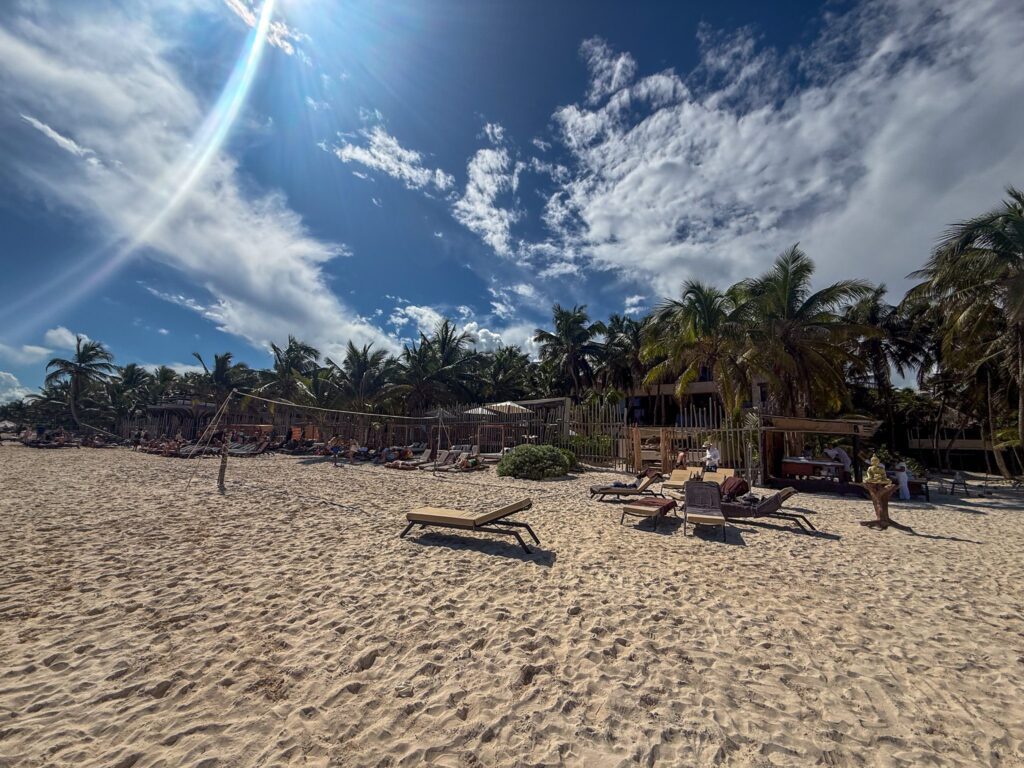 Wide sandy beach with loungers, volleyball net, palm trees, and guests relaxing under a bright sky at Secrets Tulum.