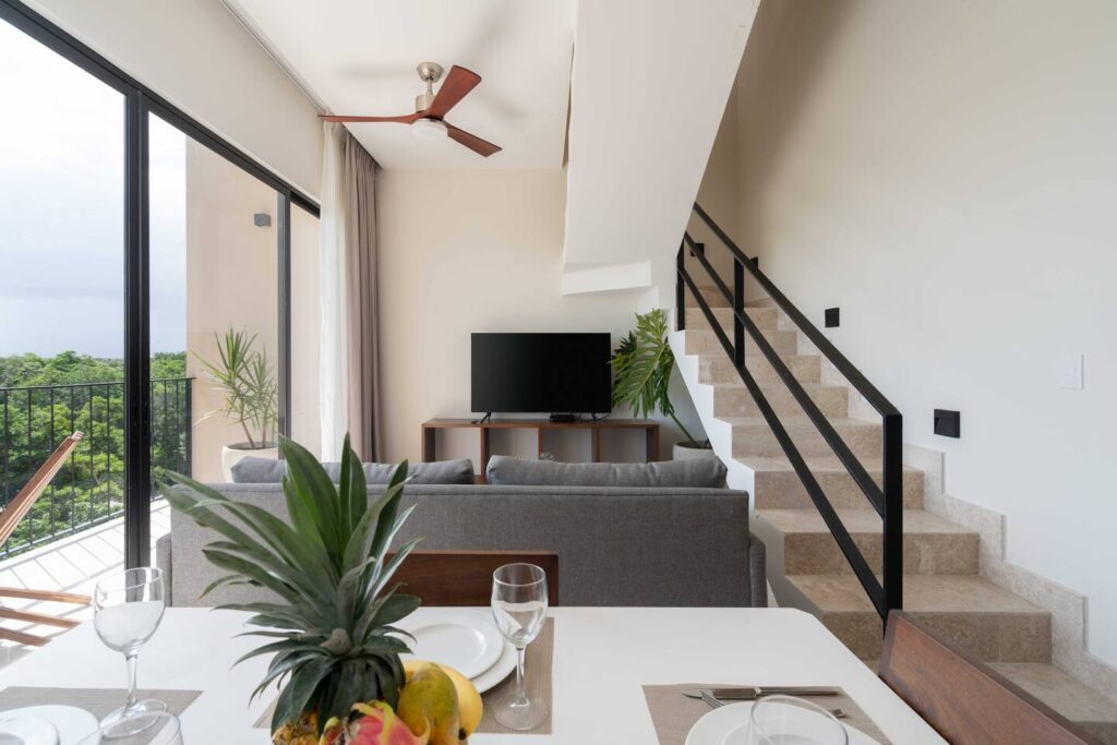 A minimalist living room with a gray sofa facing a TV, a staircase with black metal railings leading to the upper level, and large sliding glass doors that open to a balcony with a view of green trees.