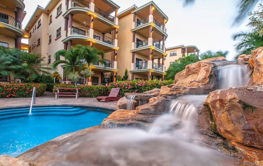 Resort view at Clarion Suites Roatán with a small pool, cascading rock waterfall, lush tropical landscaping, and multi-story hotel balconies in the background