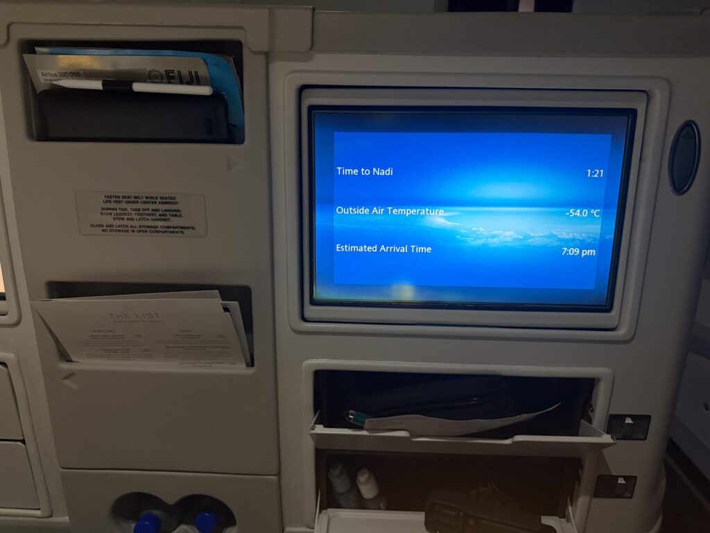 Fiji Airways A330 business class entertainment screen showing time to Nadi with visible seat storage compartments and limited personal space.