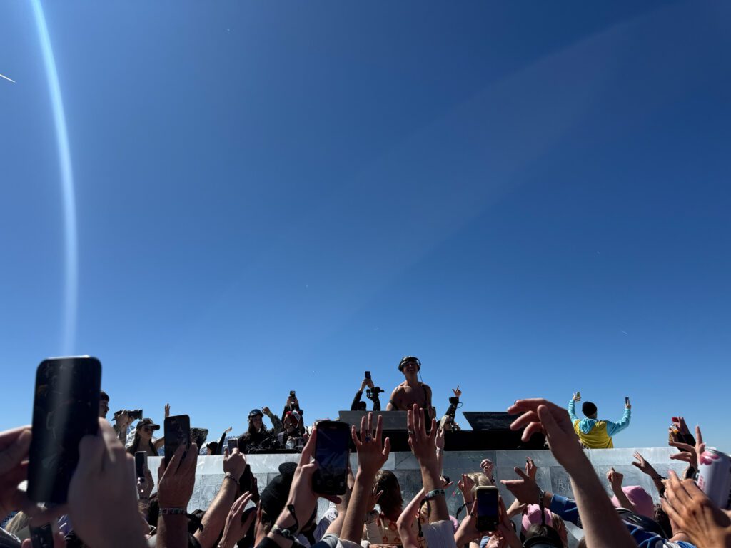 John Summit performing at Experts Only music festival with fans gathered around the DJ booth under a clear blue sky