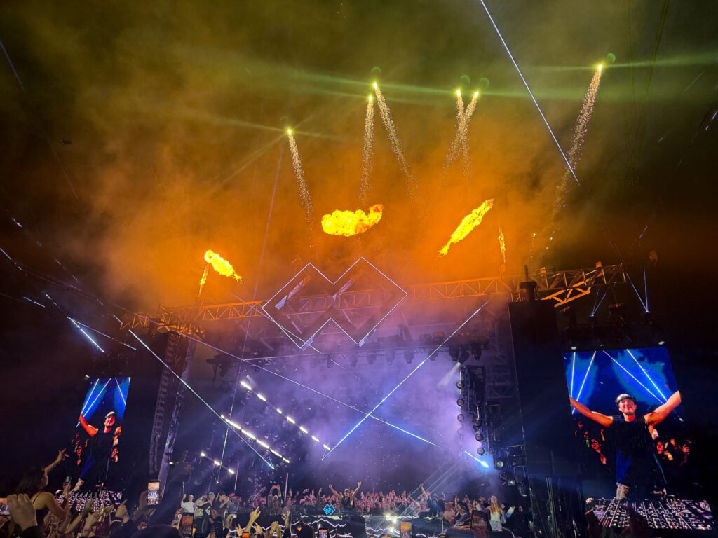John Summit performing on stage with pyrotechnics, lasers, and a packed crowd at Experts Only festival